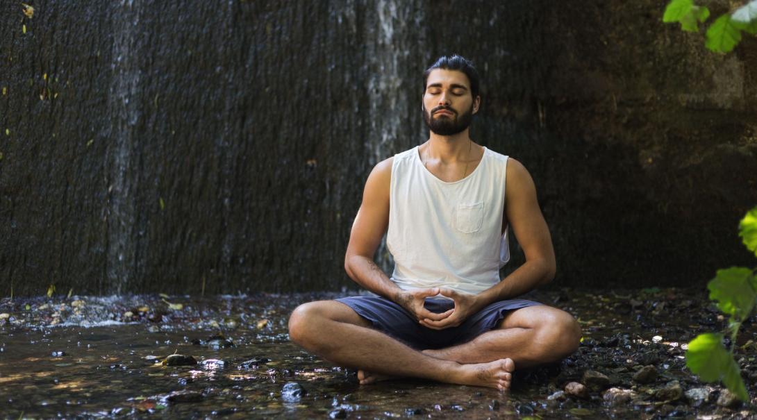 A lone man sits cross legged with his eyes closed on the ground with a small waterfall behind him