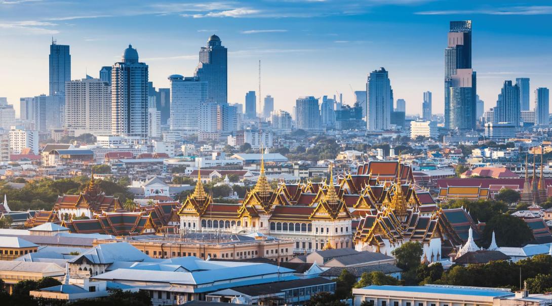 A daytime shot of Bangkok with a large, decorative, traditional building in the foreground and several modern skyscrapers lining the background 